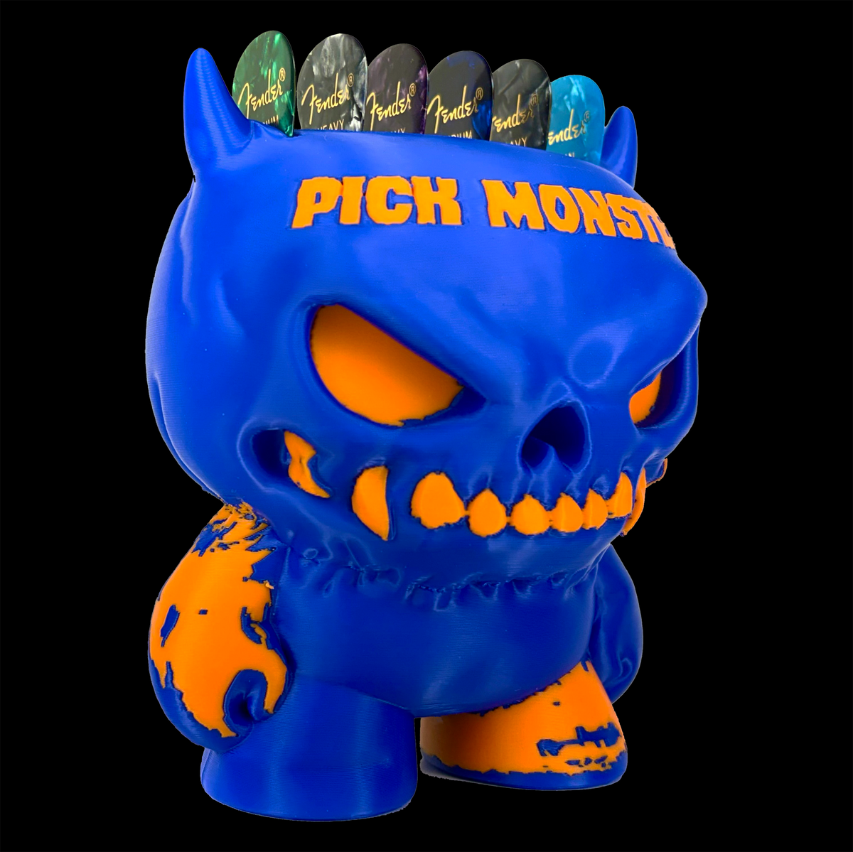 A blue and orange stylized monster figure guitar pick holder with the words “PICK MONSTERS” on its forehead, shown against a solid black background. Guitar picks are visible on its head.